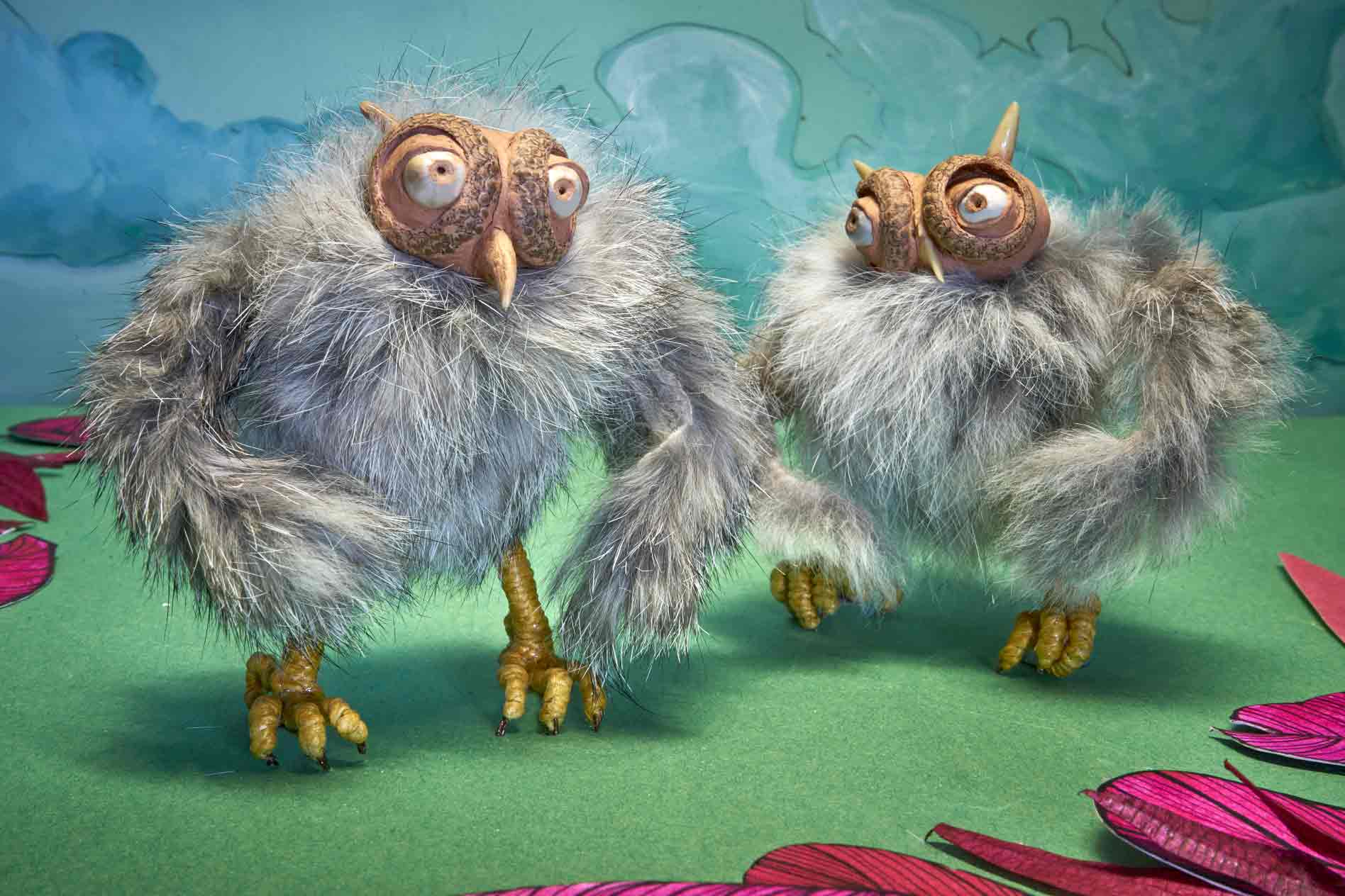 A cute pair of owl stop motion characters stand in a clearing surrounded by pink petals and a blue sky with fluffy clouds